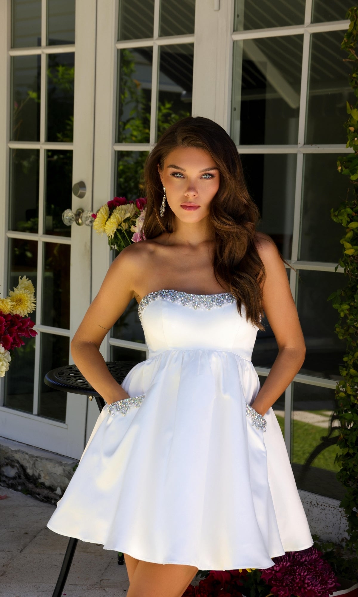 A woman in the Ava Presley 29837 strapless short hoco dress with jeweled trim stands outdoors by a glass door, surrounded by flowers and greenery.